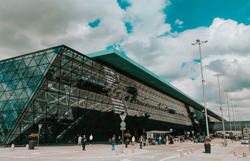 Modern glass terminal with a triangular panel facade and people walking outside on a cloudy day