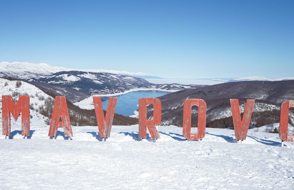 Red MAVROVO letters standing in snow with a wintry mountain landscape and a frozen lake in the background, sunny blue sky above.