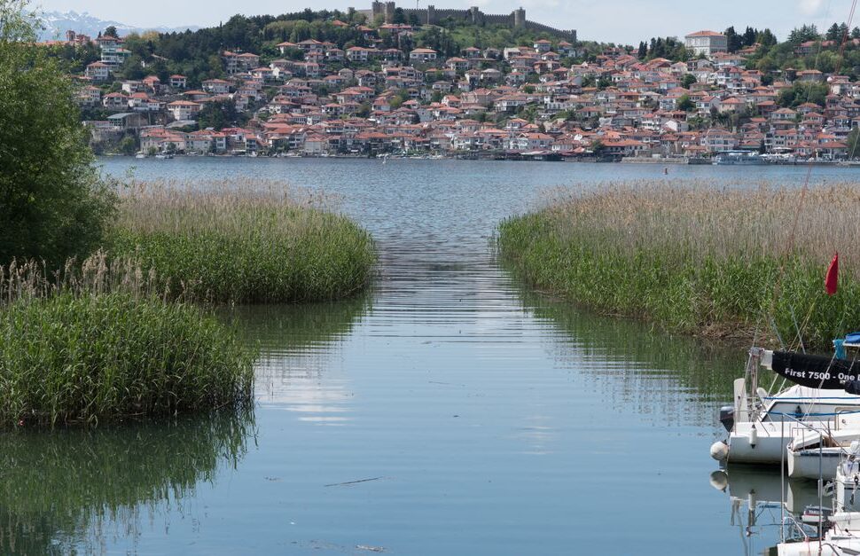 A calm lake with a narrow reed-lined channel leading toward a hillside town across the water, with small boats docked on the right.