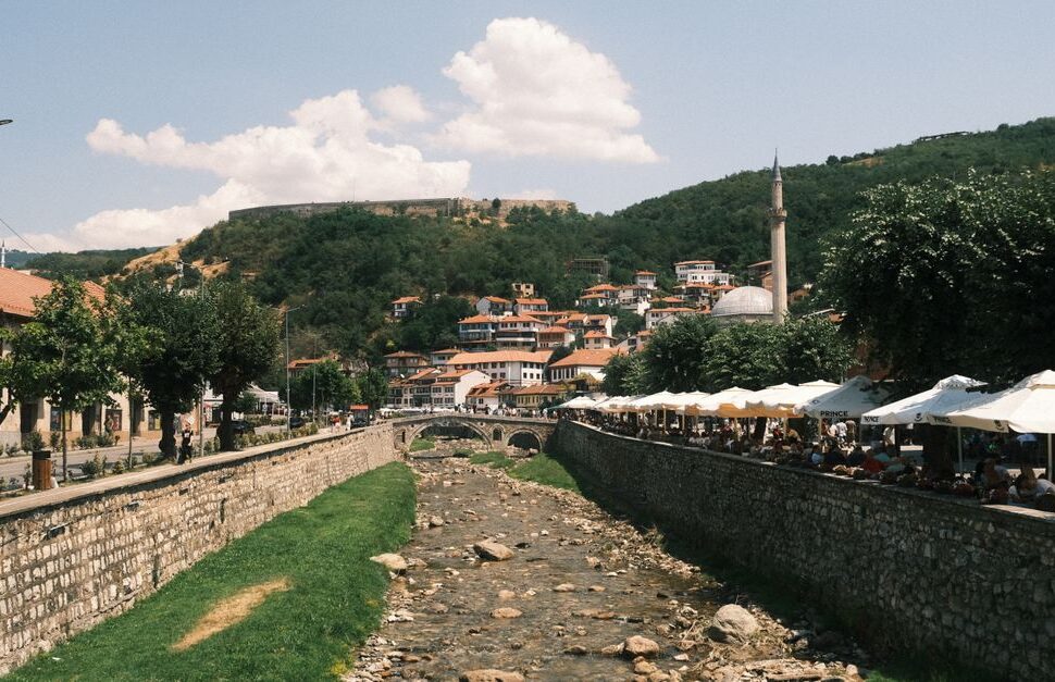 Riverside town scene with a stone embankment, a rocky riverbed, a stone bridge, and a riverside cafe shaded by white umbrellas on the right; hillside houses in the background.