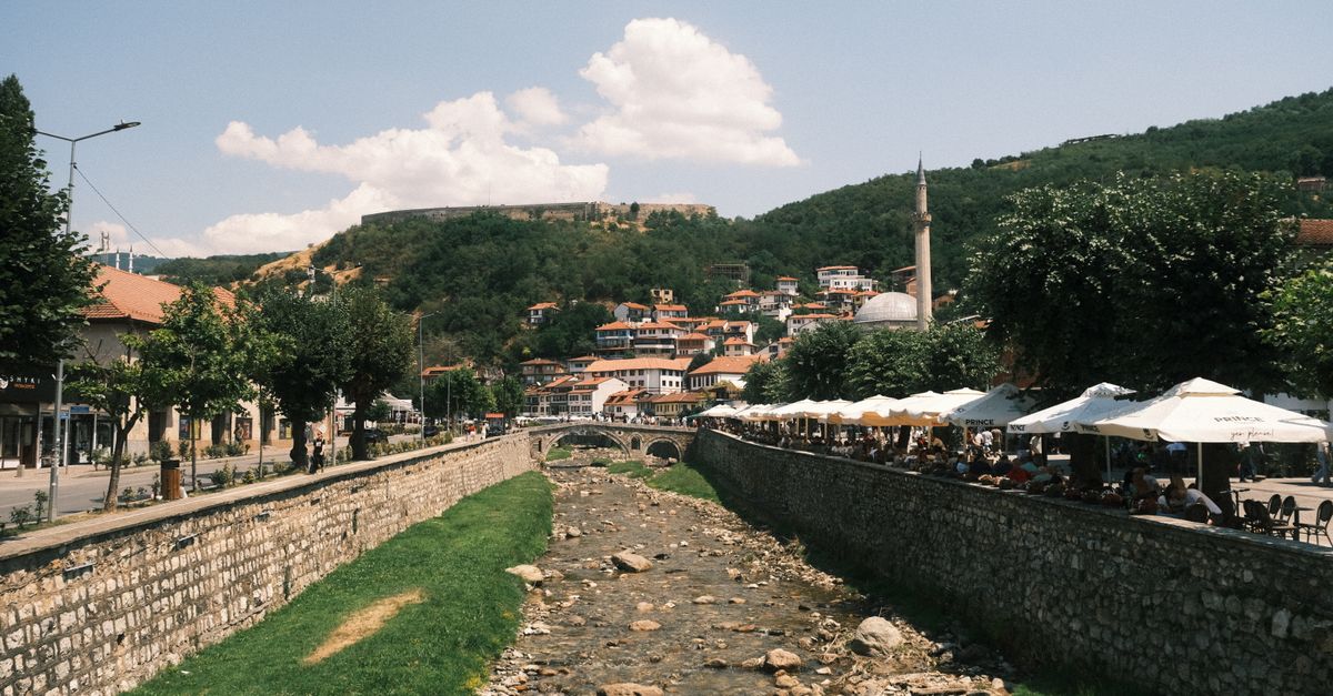 Riverside town scene with a stone embankment, a rocky riverbed, a stone bridge, and a riverside cafe shaded by white umbrellas on the right; hillside houses in the background.
