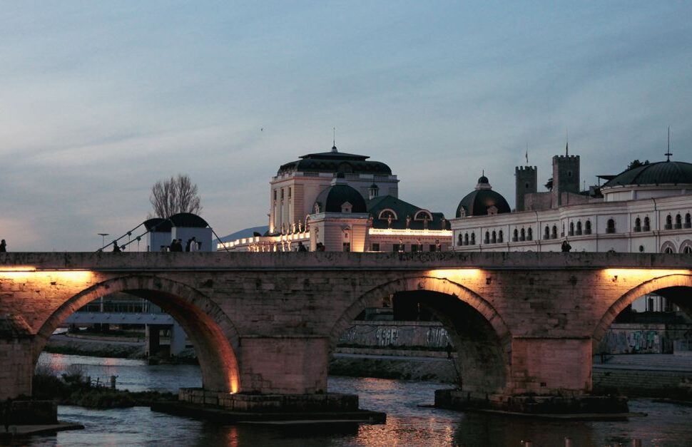 Stone arch bridge over a calm river at dusk, warm lights under the arches, with a domed historic building behind.
