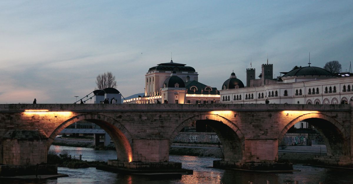 Stone arch bridge over a calm river at dusk, warm lights under the arches, with a domed historic building behind.