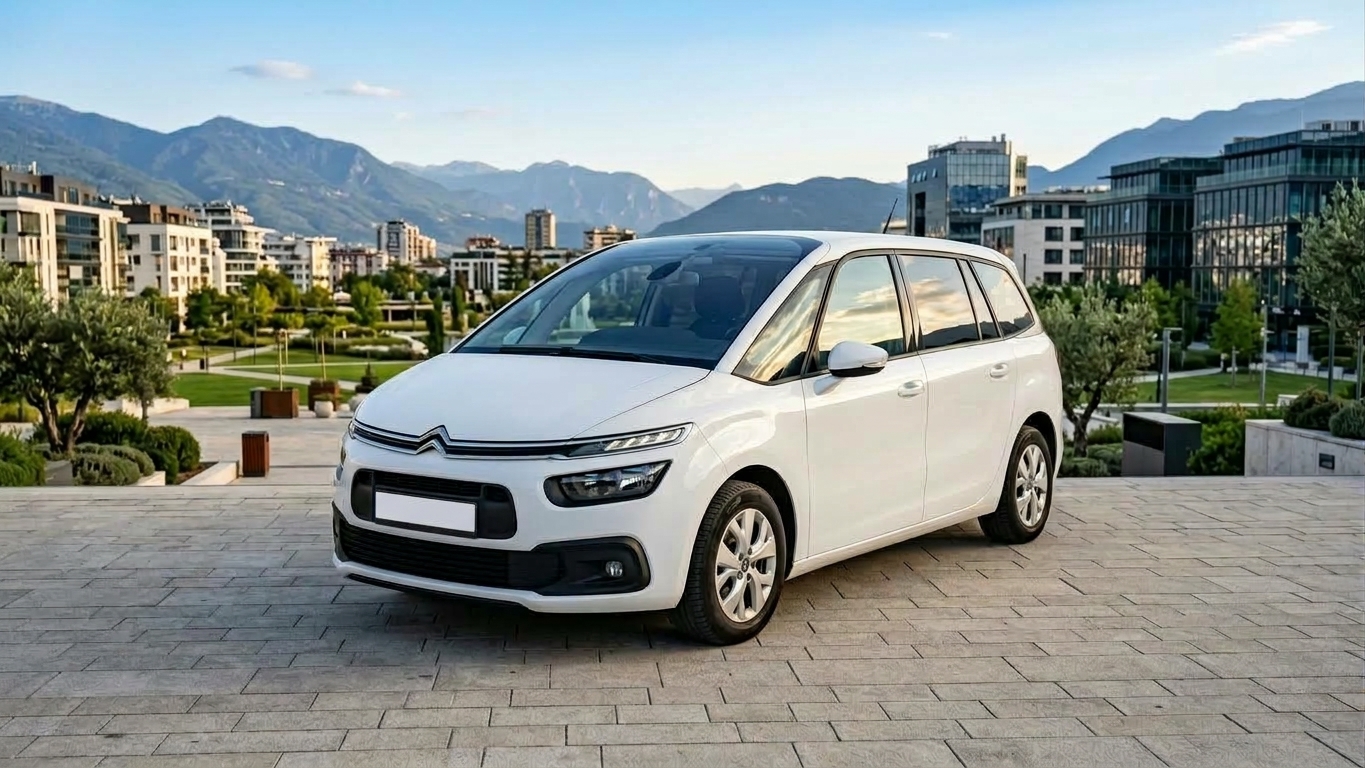 White multi-seat car parked on a stone plaza with modern buildings and green hills in the background.